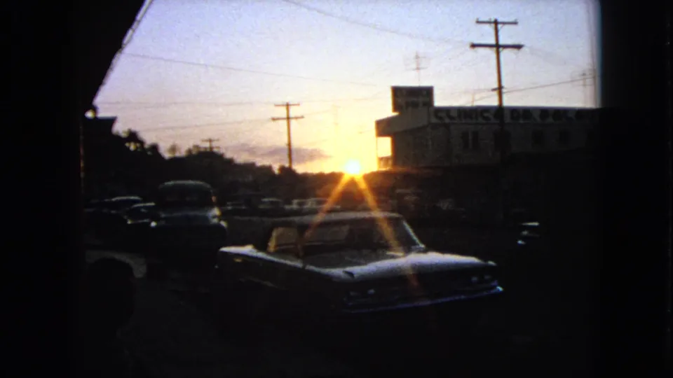 1963 8mm street view with a parked classic car, strong sun flare, utility poles, and a silhouetted commercial building.