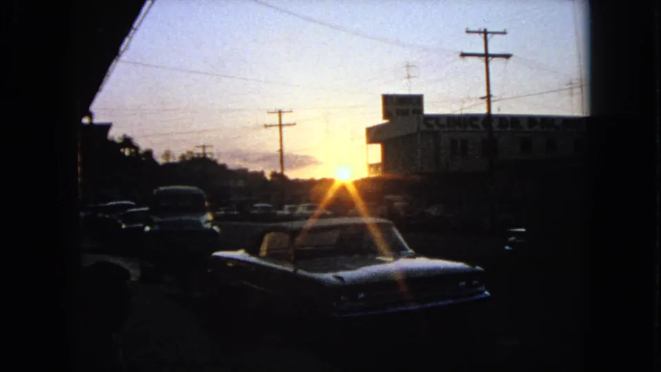 1963 Barrington, Illinois dusk frame showing parked cars, a clinic exterior, overhead wires, and strong sun flare.