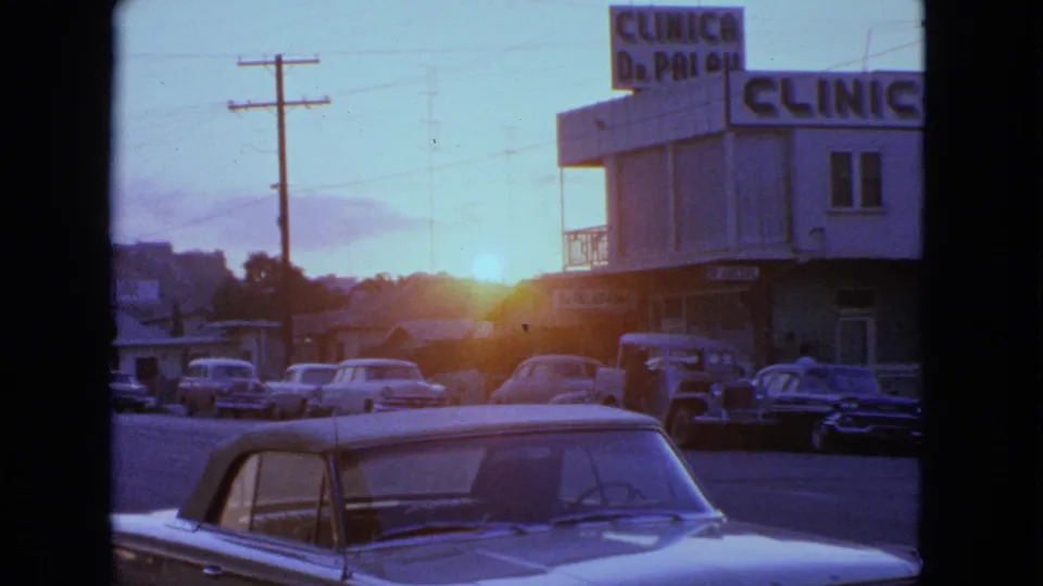 1963 Barrington street view with parked cars, low sun flare, utility poles, and a building marked with clinic signage.