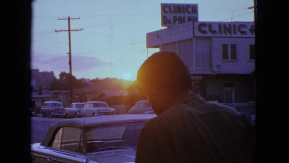 A silhouetted man crosses the foreground of a 1963 Barrington street scene with parked cars, sunset flare, and clinic signage.