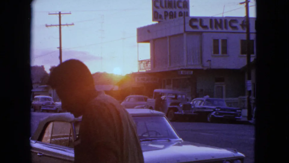 The man continues across the foreground, with 1960s cars, a clinic sign, utility poles, and warm sun flare behind him.
