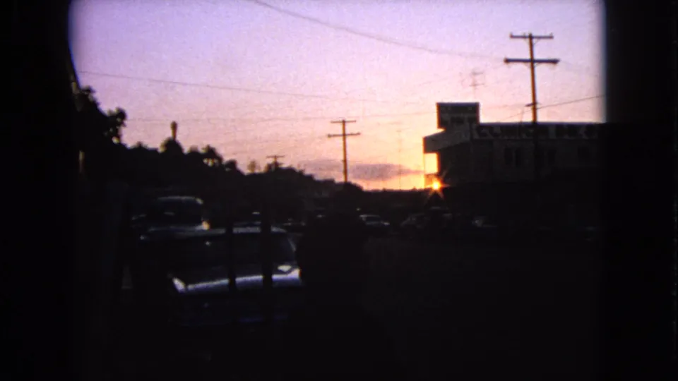 1963 Barrington street scene with cars in deep shadow, utility poles, overhead wires, and a sunset glow beside a low commercial building.