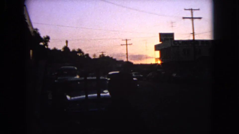 A final 1963 Barrington dusk frame showing parked cars, utility poles, wires, trees, and a low building lit from behind by sunset.