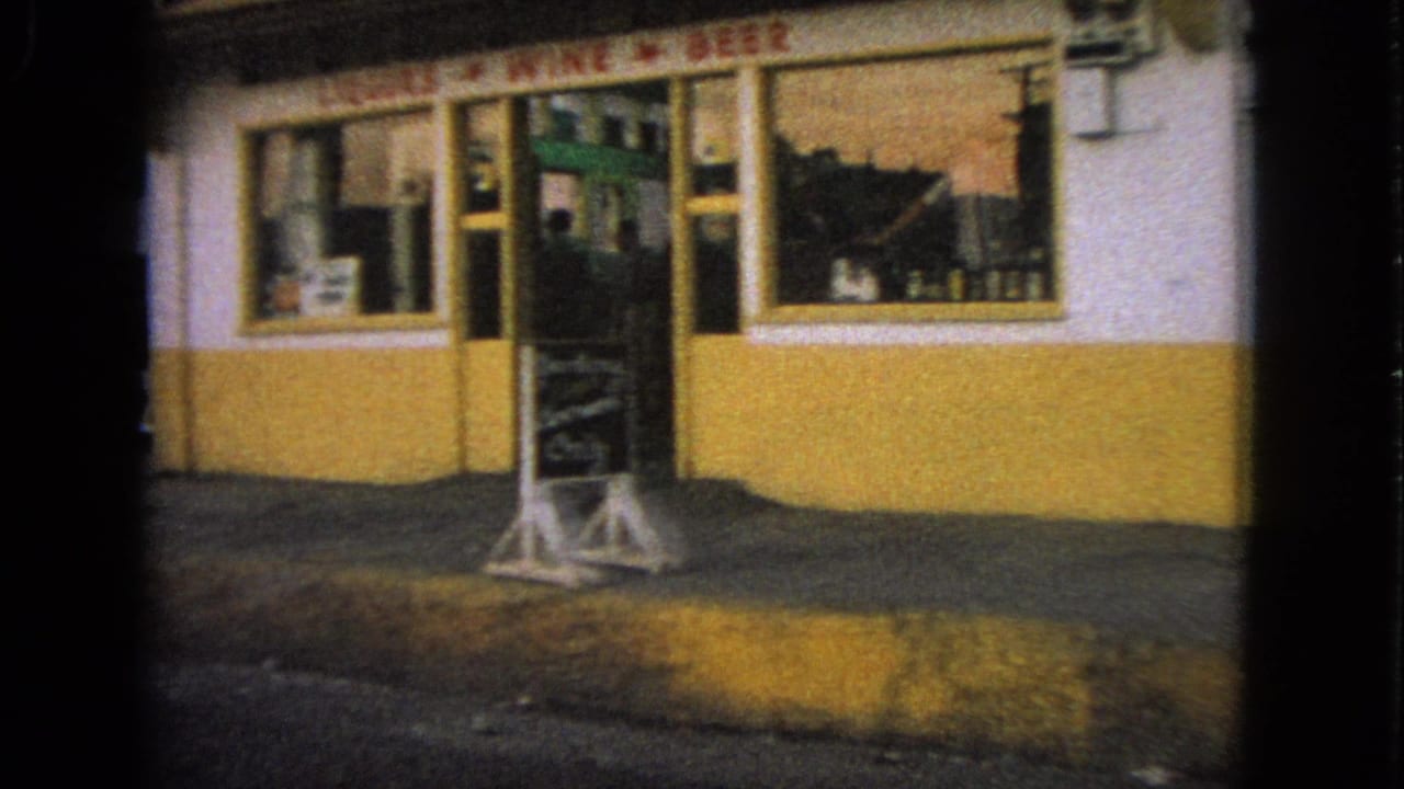 1963 home-movie view of a small liquor store exterior in Barrington, Illinois, with window signage and a freestanding sidewalk board.