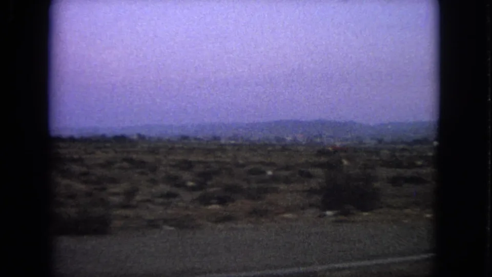 Wide home-movie landscape view, likely from travel footage, showing dry ground, distant hills, and a purple-shifted sky.