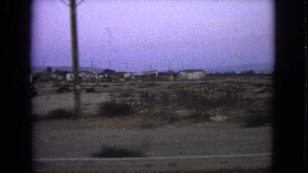Barrington-context 1963 home-movie frame with a foreground utility pole, sparse roadside land, distant buildings, and soft 8mm color shift.