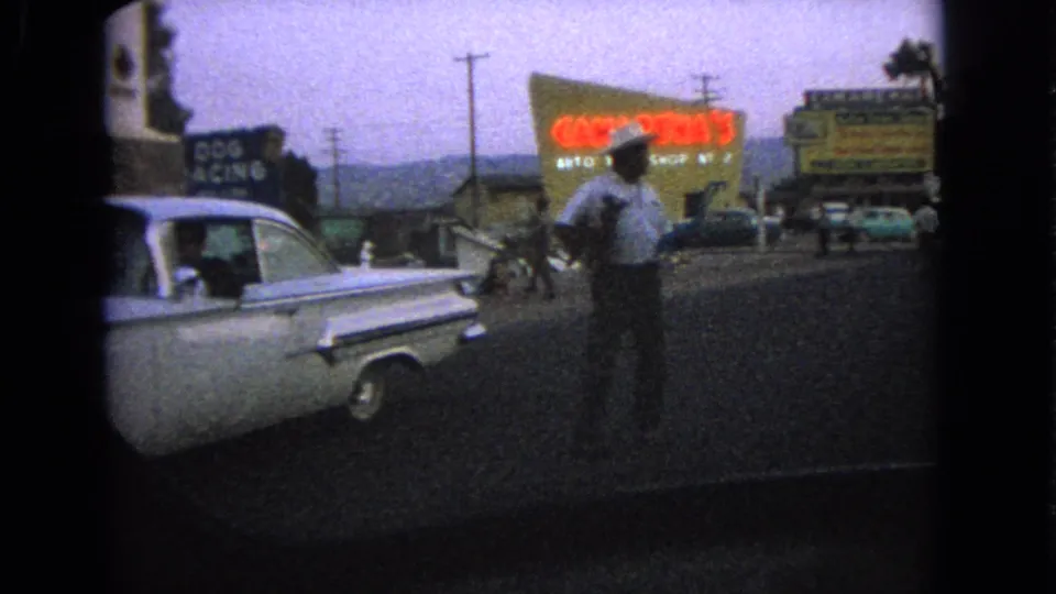 1963 home-movie frame showing a man near a mid-century car, with colorful roadside signs or billboards and buildings in the background.