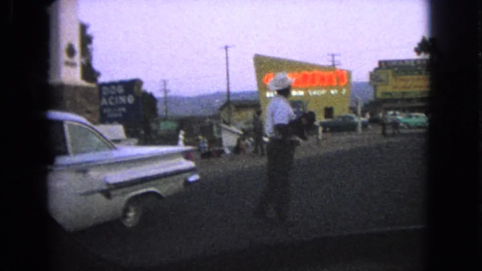 1963 Barrington-context roadside frame with a man by a classic car and bright visible signage or billboards behind him.