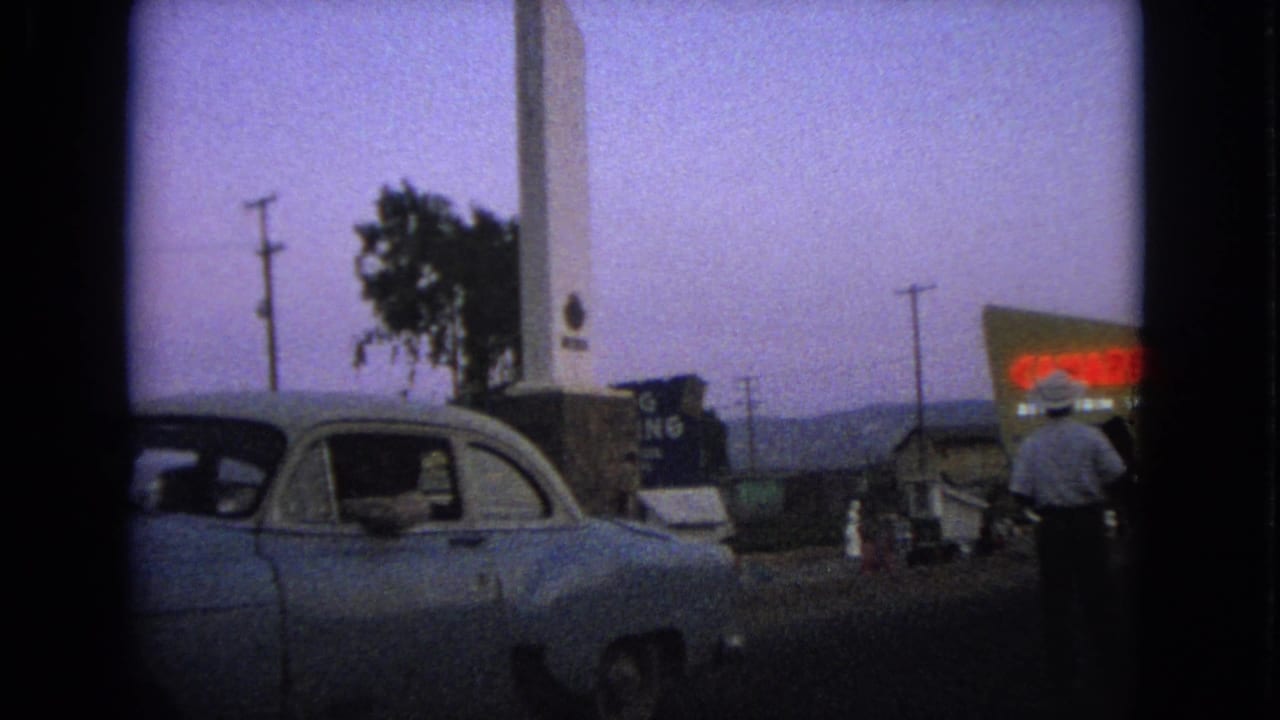 1963 Barrington, Illinois home-movie frame showing a parked or stopped classic car, a tall white monument, roadside signs, and a pedestrian.