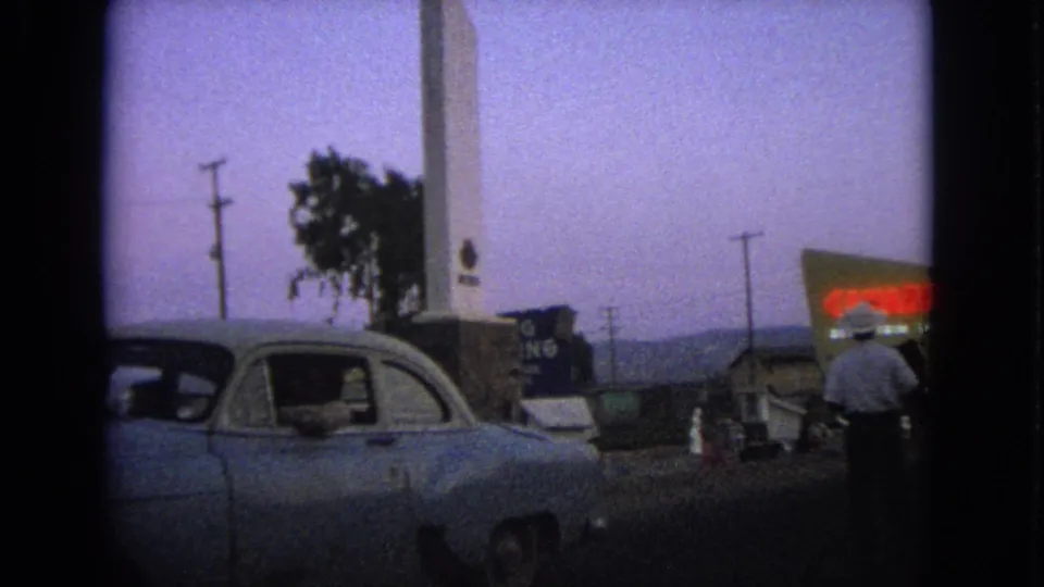 1963 Barrington, Illinois home-movie frame showing a parked or stopped classic car, a tall white monument, roadside signs, and a pedestrian.