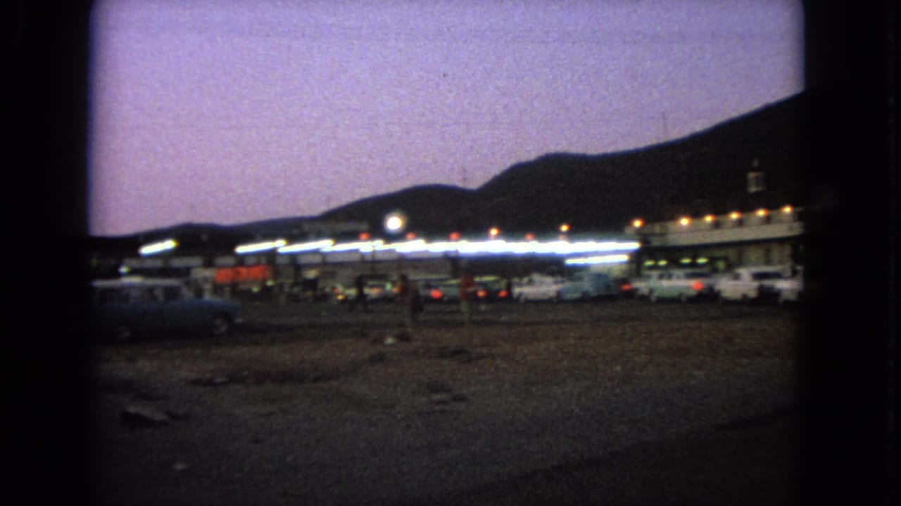 Wide 1963 Barrington drive-in view with glowing building lights, parked cars, rough lot ground, and hills under a purple sky.