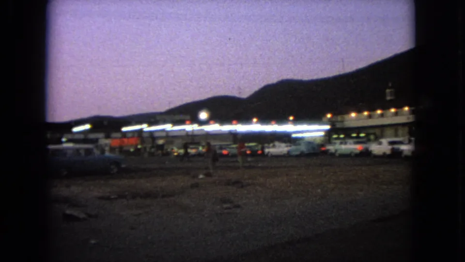 Wide 1963 Barrington drive-in view with glowing building lights, parked cars, rough lot ground, and hills under a purple sky.