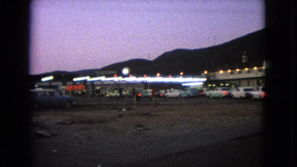 Barrington drive-in scene from 1963 showing a lit concession or theater area, parked cars, small patrons, and dark hills at dusk.