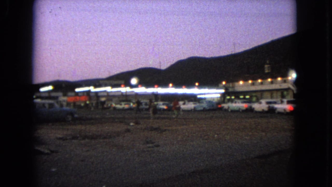 1963 Barrington, Illinois 8mm frame showing parked cars near a lit drive-in or roadside building at dusk.