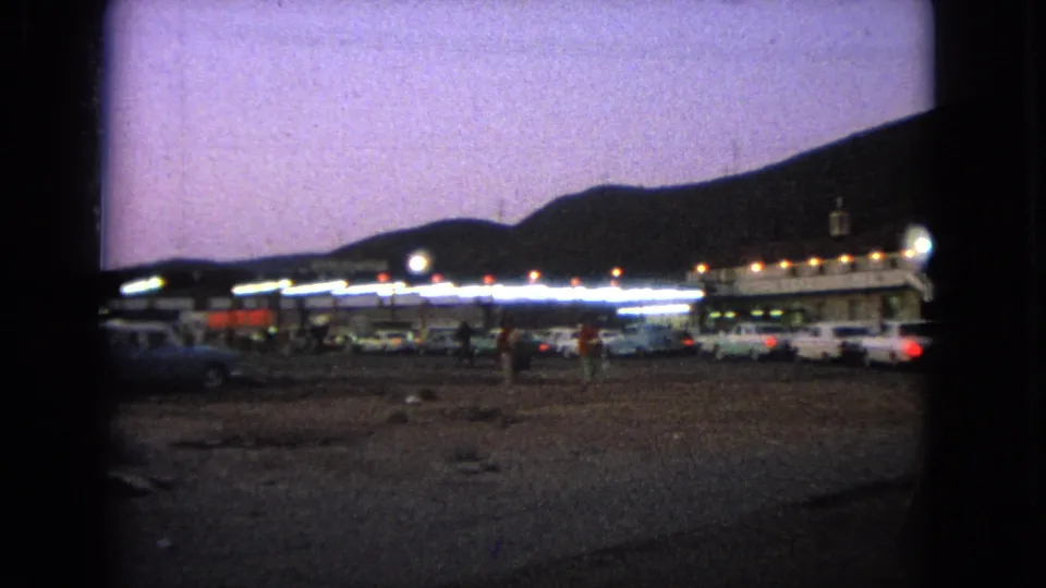 1963 Barrington, Illinois frame with distant people among parked cars outside a lit drive-in or roadside entertainment area.