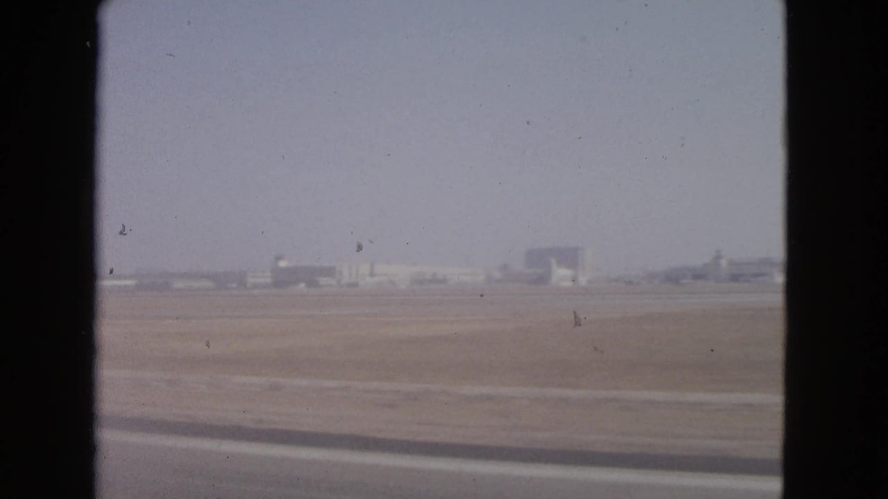 Hazy airport field seen through a dark window edge with distant low buildings.