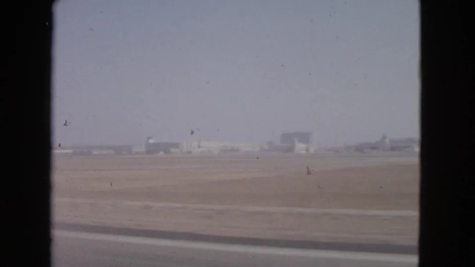 1963 Barrington, Illinois airport field viewed through a dark window edge, with runway pavement and distant buildings under a pale sky.
