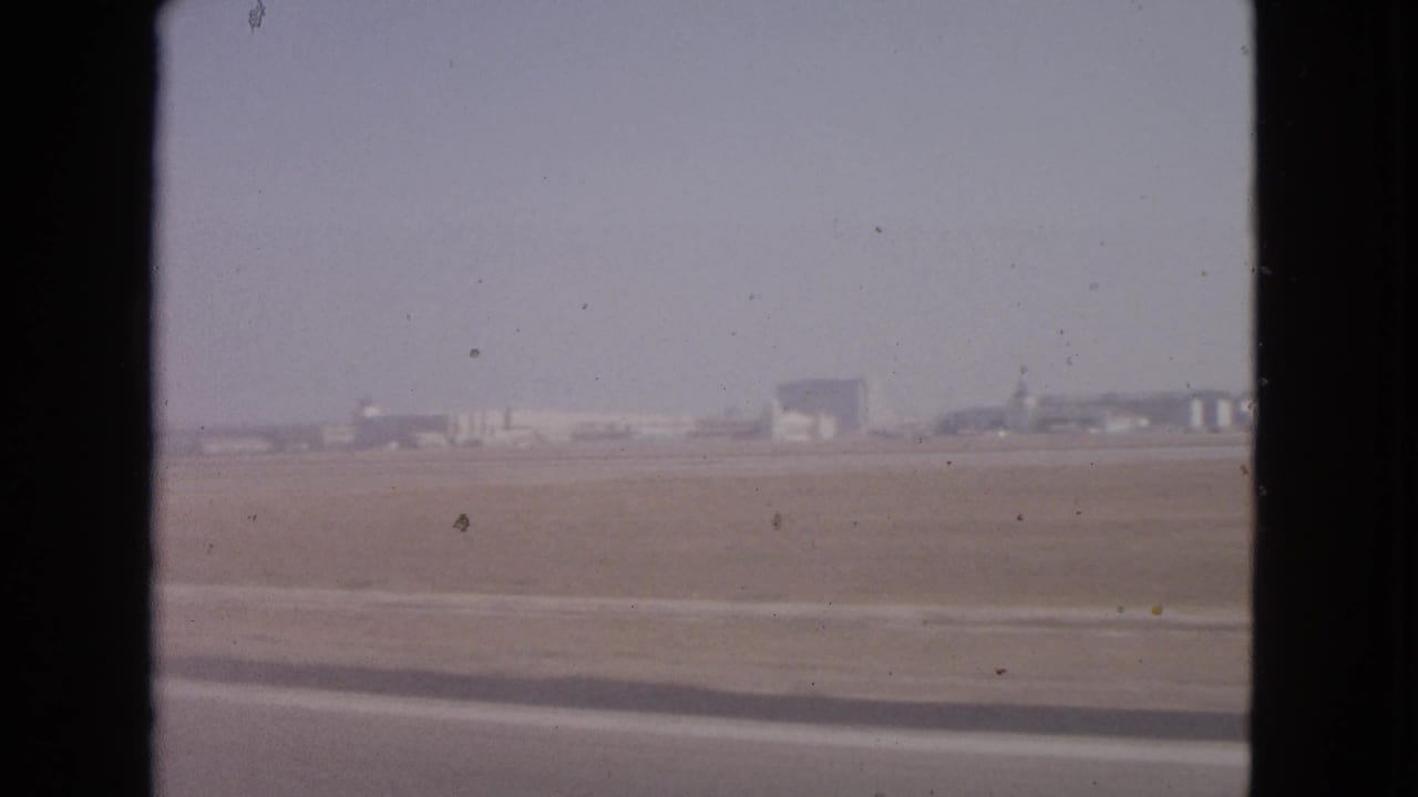 A moving 8mm view of Barrington airport grounds in 1963, with distant hangar-like buildings and a broad runway area.