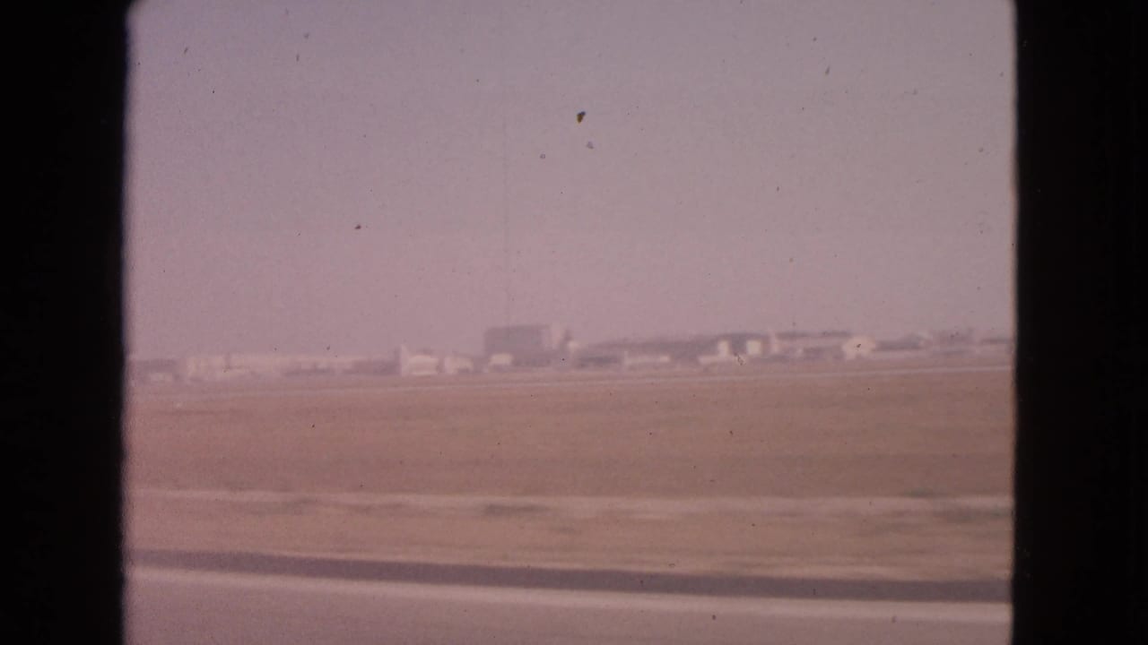 Flat roadside landscape seen through a car window with distant low buildings on the horizon.