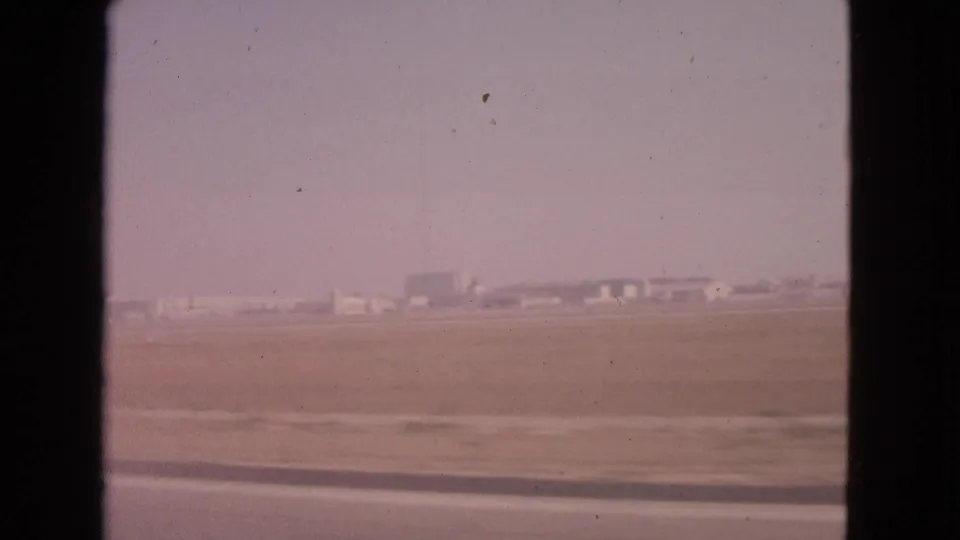 1963 Barrington, Illinois 8mm frame showing open flat ground, a roadside strip, and distant low structures through a moving car window.
