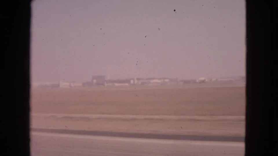 1963 8mm travel frame near Barrington, Illinois showing dry open ground, a road edge, and distant low structures under a pale sky.