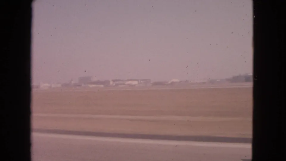 Barrington, Illinois 1963 home-movie still with flat roadside terrain, distant low buildings, and a dark car-window divider in the frame.