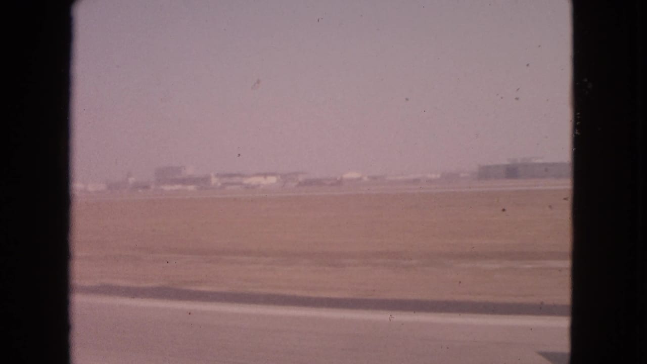 1963 8mm passenger-window view over a flat airfield near Barrington, Illinois, with runway pavement and distant buildings.