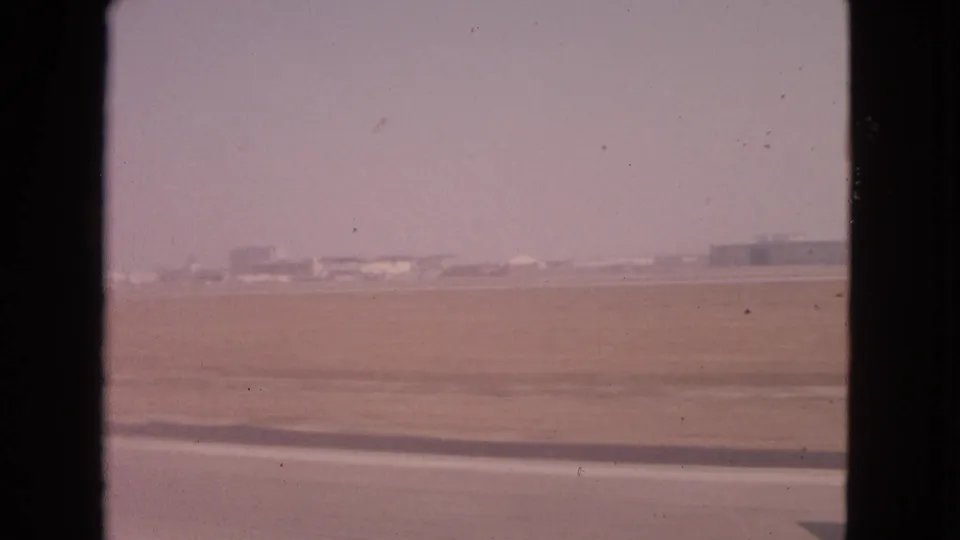 1963 8mm passenger-window view over a flat airfield near Barrington, Illinois, with runway pavement and distant buildings.