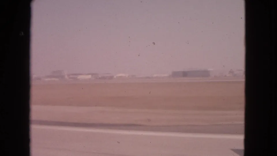 Passenger-window view of a hazy 1963 airfield with low buildings and runway surfaces visible beyond the glass.