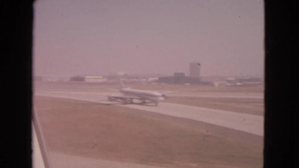 1963 8mm view from an aircraft window showing a distant plane moving along the runway at an airport.