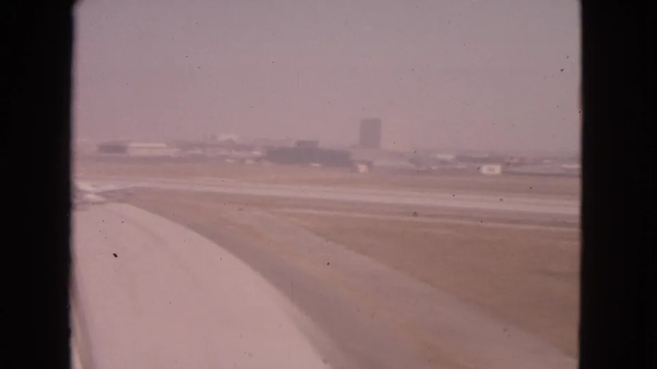 Passenger-window view of an aircraft wing over runway pavement and distant airport buildings in 1963 Illinois footage.