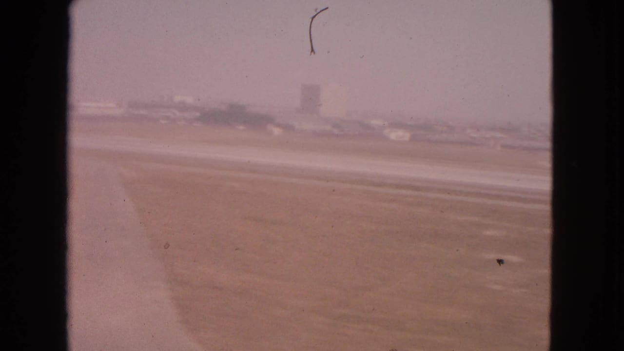Hazy airport runway seen through dark window-frame edges with distant buildings and aircraft shapes.