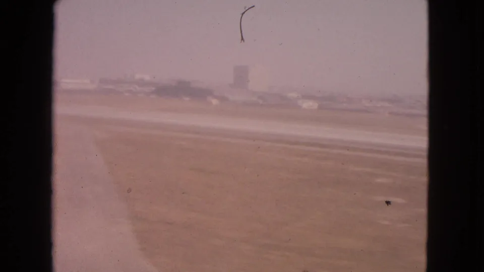 1963 Barrington, Illinois airport view through a window, showing runway pavement, haze, distant airport buildings, and small aircraft forms.