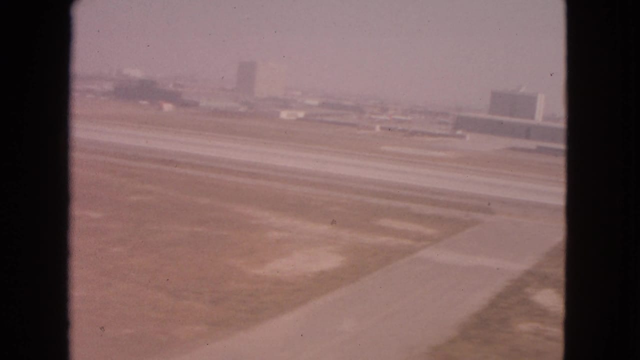 Airport runway and taxiway with low buildings in the distance, framed by dark vertical window edges.