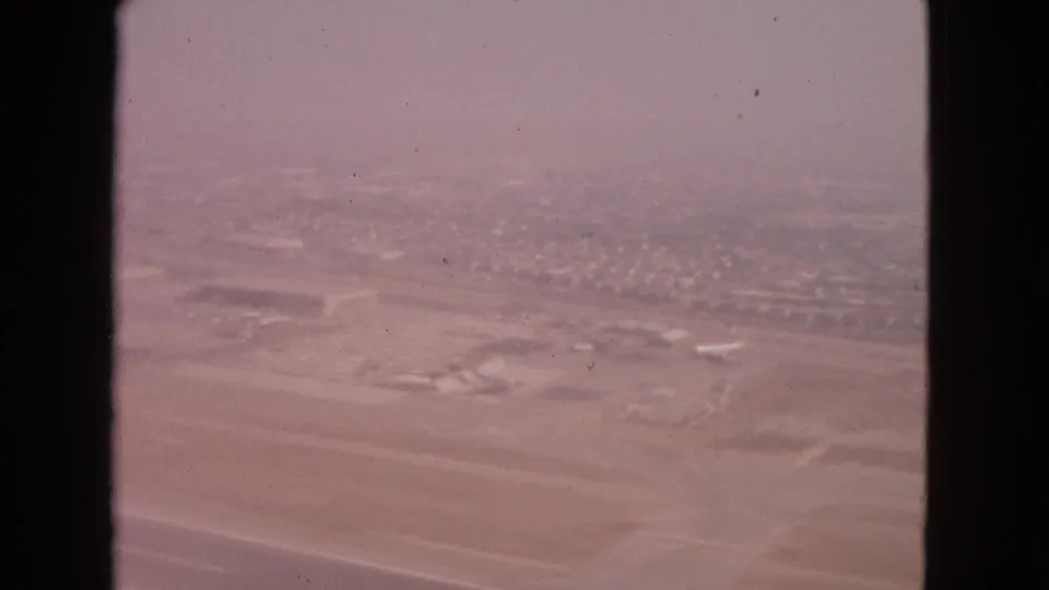 1963 Barrington, Illinois aerial home-movie frame showing airport grounds, small aircraft, and neighborhoods beyond in soft haze.
