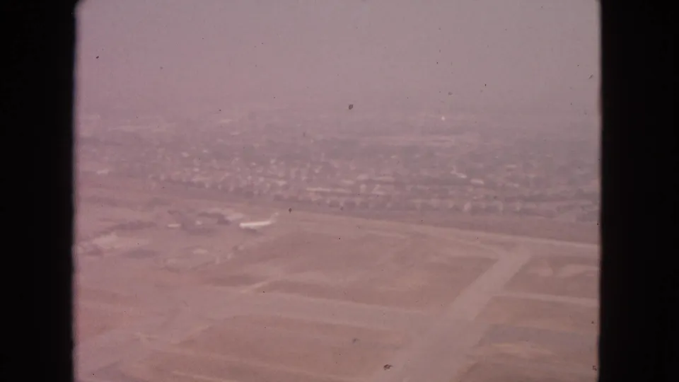 1963 home-movie aerial frame over Barrington, Illinois, showing intersecting runway pavement, open airport land, and suburban housing.