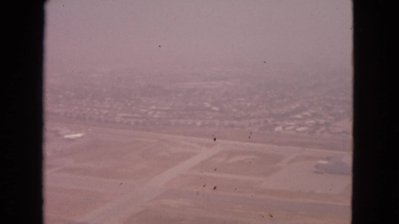 1963 8mm airplane-window view over an airport area near Barrington, Illinois, with runways, open ground, and distant development softened by haze.