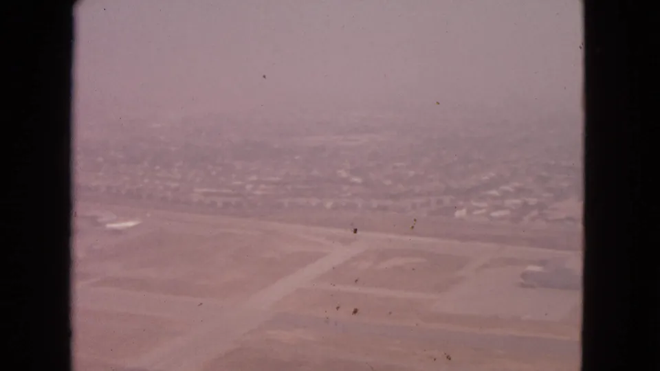 1963 8mm airplane-window view over an airport area near Barrington, Illinois, with runways, open ground, and distant development softened by haze.