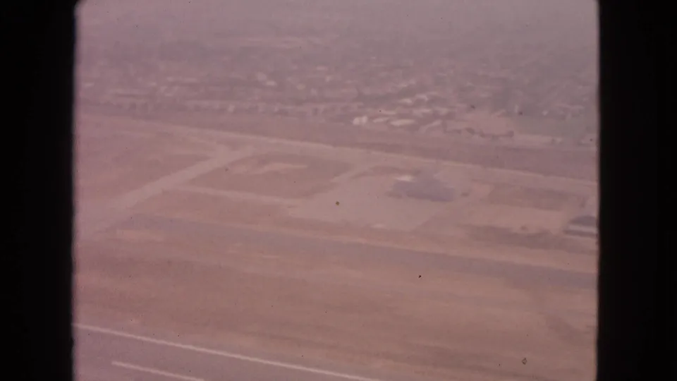 Oblique 1963 aerial view from a plane window showing runway geometry, open airport land, and distant neighborhoods in a hazy landscape.