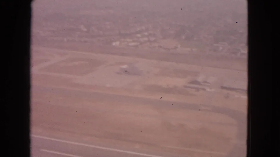 1963 passenger-window aerial over airport grounds, with runway markings, open fields, and small low structures visible through soft 8mm color.