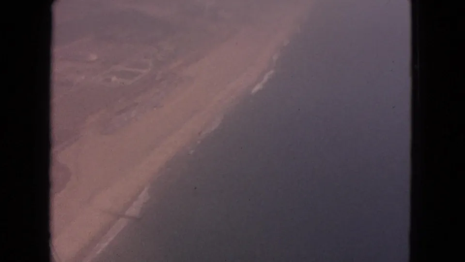 Passenger-window aerial from 1963 showing a broad water edge or shoreline beside open land, with the aircraft frame visible at the side.