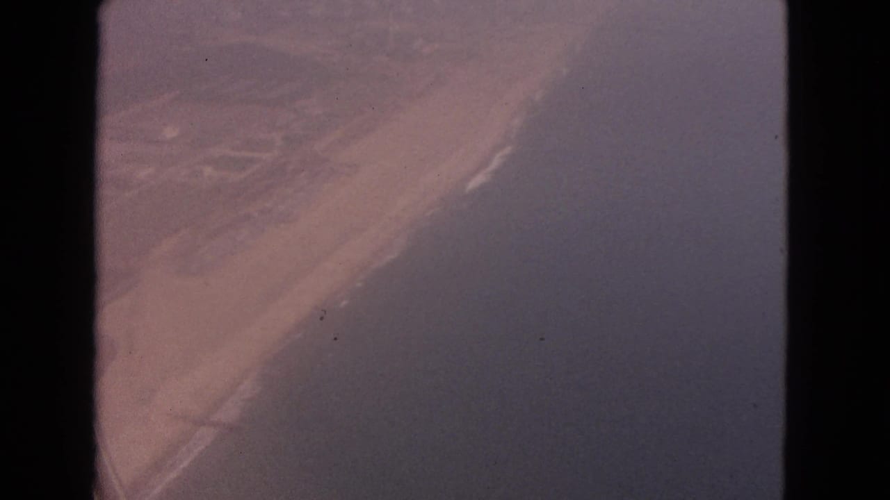 1963 8mm aerial view of a diagonal sandy shoreline with surf and darker water, location not identifiable in-frame.