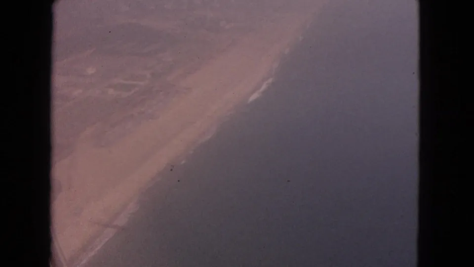 1963 8mm aerial view of a diagonal sandy shoreline with surf and darker water, location not identifiable in-frame.
