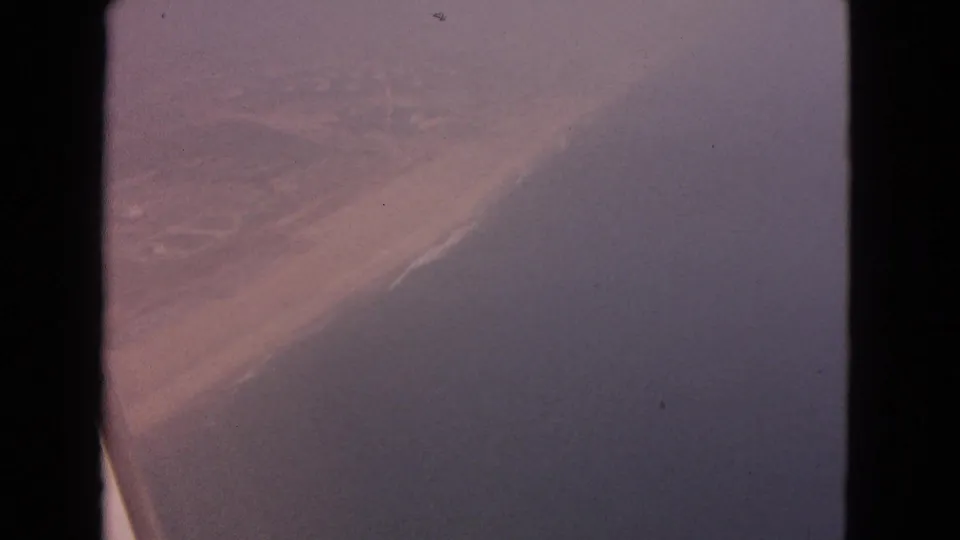 1963 home-movie aerial view of a pale sandy shore and darker water with soft 8mm grain and color shift.