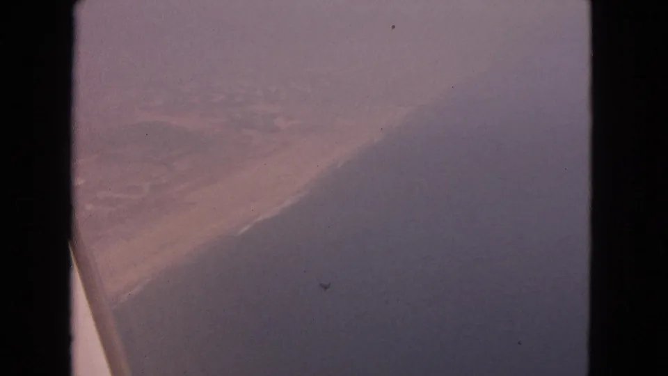 Muted 1963 8mm aerial shoreline frame showing surf along sand, with no people or landmarks visible.