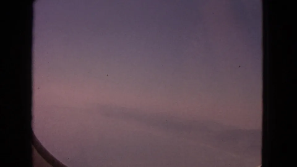 1963 8mm home-movie view from an airplane window, showing a pink sunset sky with faint clouds and a dark window edge.