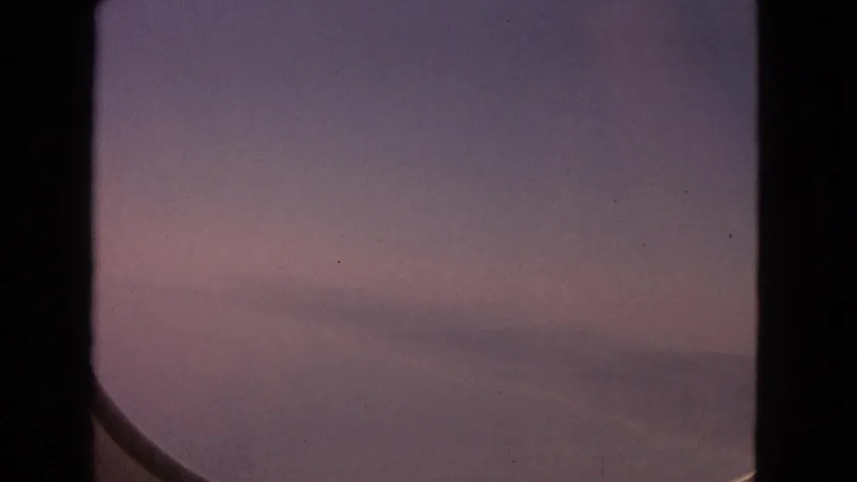 Metadata-supported 1963 aerial travel frame with a muted pink sky, low cloud band, and curved airplane window edge.