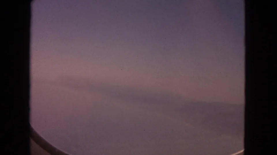 1963 8mm passenger-window view showing lavender and rose sunset tones, faint clouds, and the curved edge of the aircraft window.