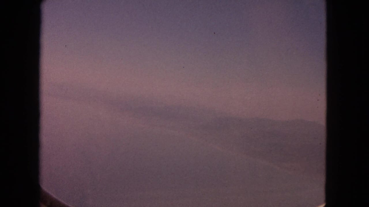 1963 8mm frame showing a hazy aerial view of distant mountains and water through a curved airplane window.
