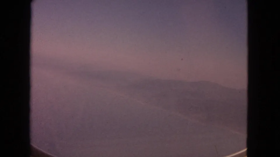 A soft 1963 8mm aerial frame with distant ridges and water partly framed by the aircraft window.